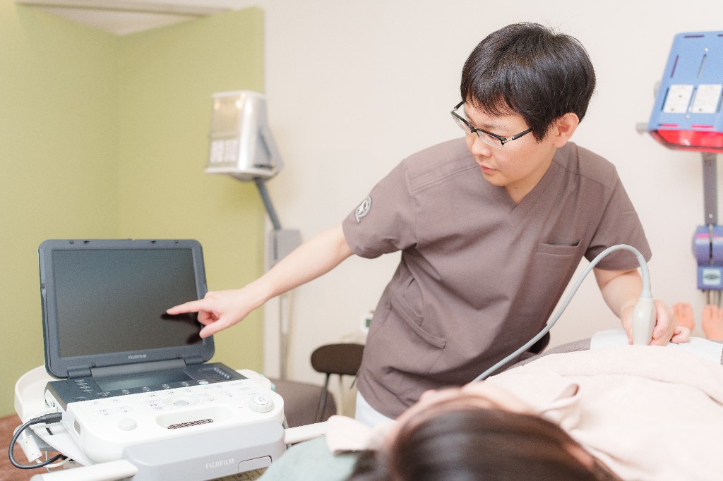 逆子鍼灸の施術イメージ。施術ベッドで腹部のエコー検査を行う施術者と、リラックスして受ける妊娠中の方。
